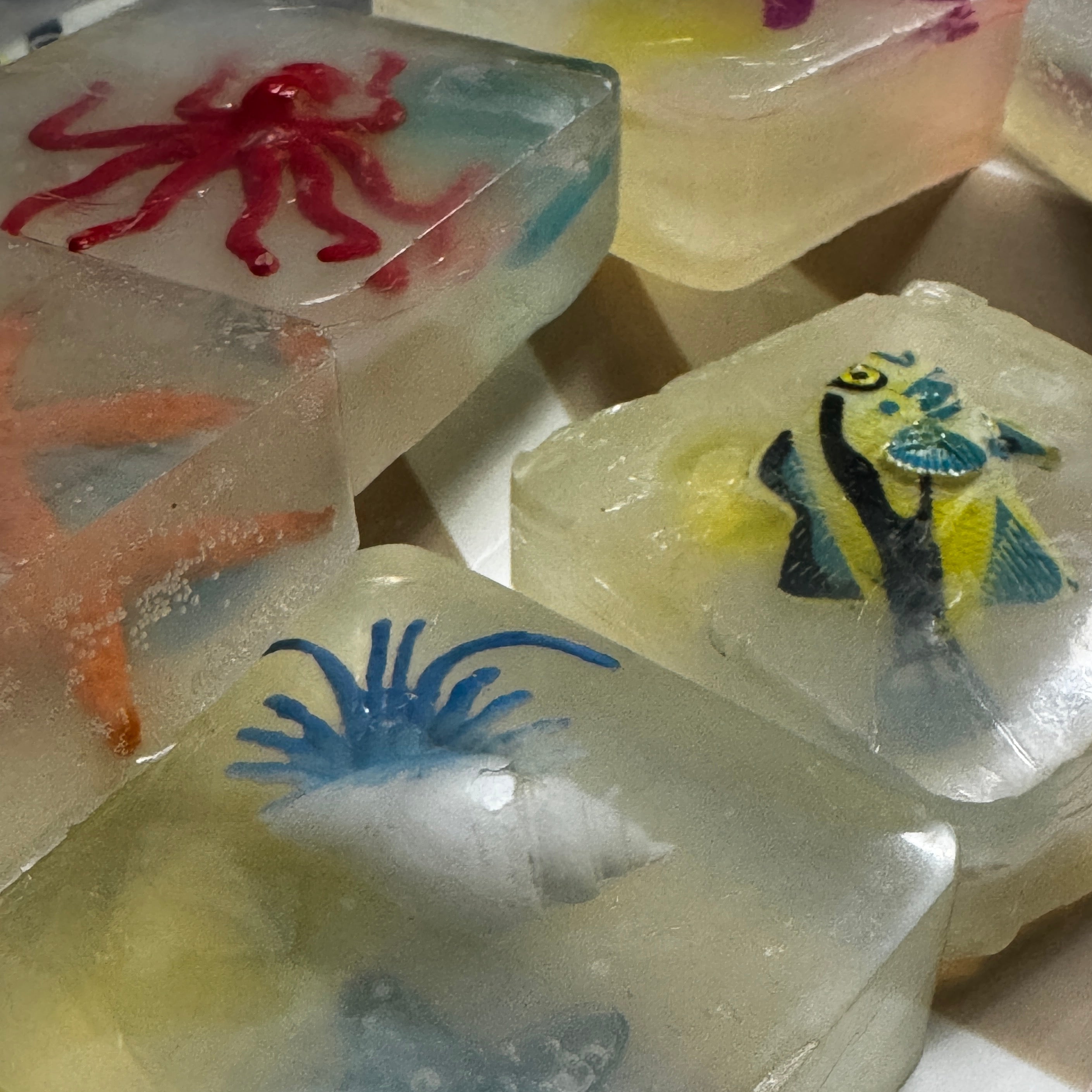 Several translucent square soaps with embedded sea-themed designs, including a red octopus, blue anemone, yellow and blue fish, and an orange starfish, arranged on a light surface.