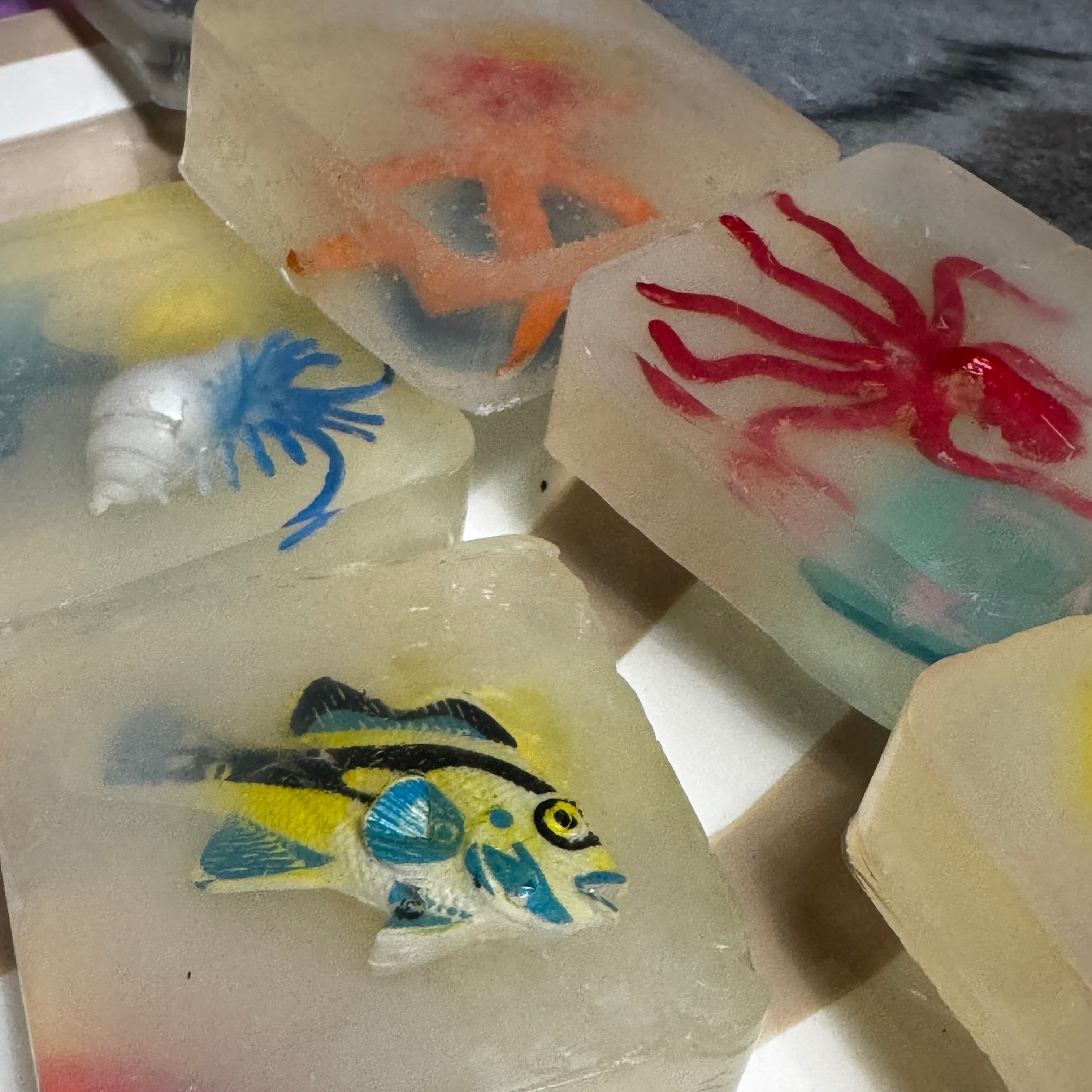 A group of translucent rectangular soap bars featuring colorful sea life designs, including a yellow and blue fish, a red spider, a blue octopus, and a white shell, arranged on a white surface.