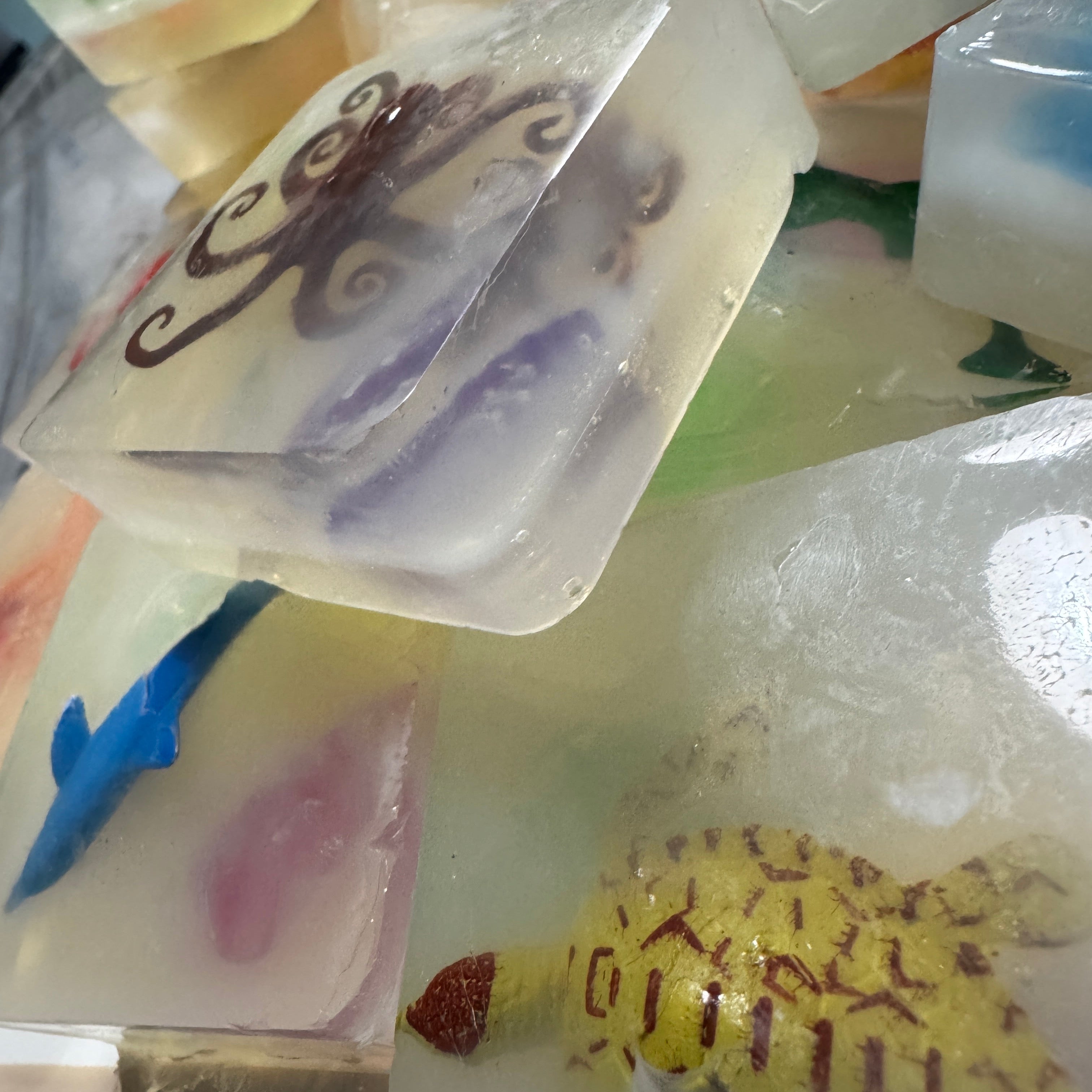 A close-up pile of translucent rectangular soap bars or wax melts, some containing embedded decorative elements like a purple octopus, a blue fish, and a yellow patterned turtle.
